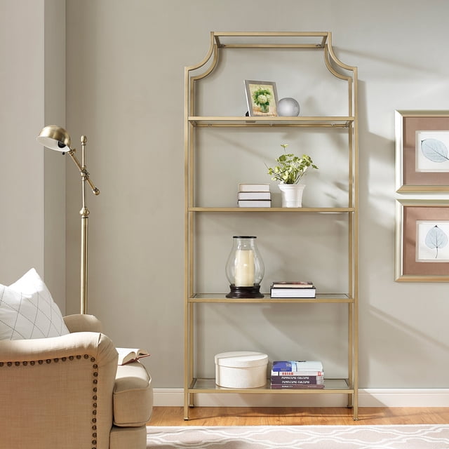 crosley aimee etagere bookcase Soft Gold pagoda-styled steel frame with Art Deco curves, centered front view with four tempered glass shelves (20-lb each) holding books, a potted plant, framed photo and decorative vase; staged beside a floor lamp and upholstered armchair, suitable for display shelving and modular combination for expanded storage.